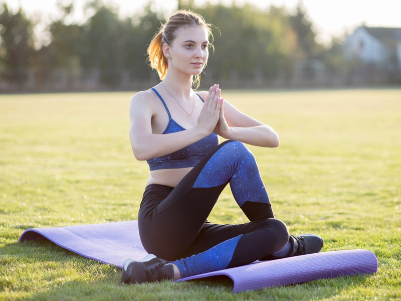 young woman performing a seated yoga pose on a purple mat outdoors in a park at sunset